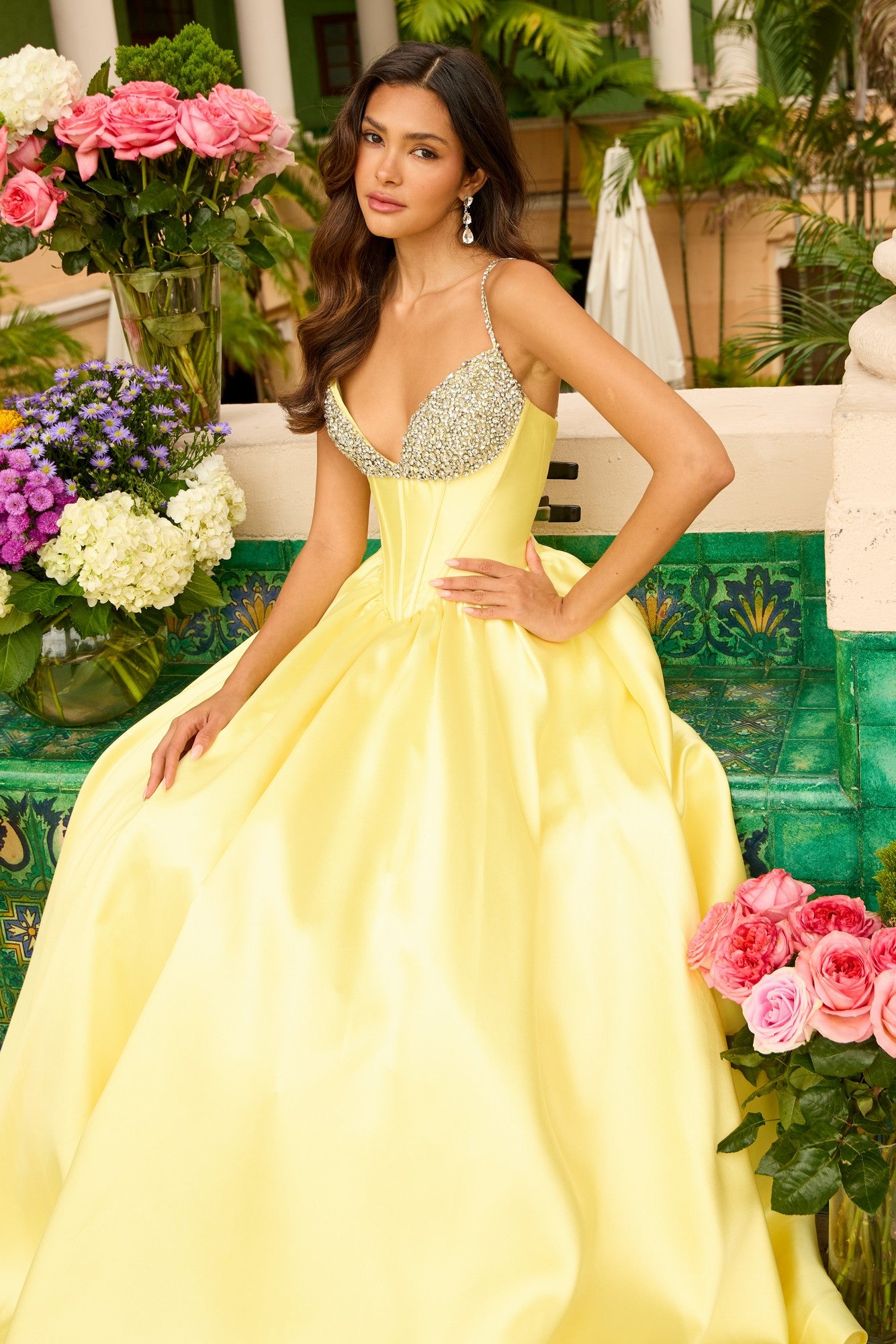 Woman in a soft yellow Ava Presley 42311 long prom dress with a jeweled corset bodice sits on a tiled bench, surrounded by vases of pink and purple flowers in an outdoor garden.